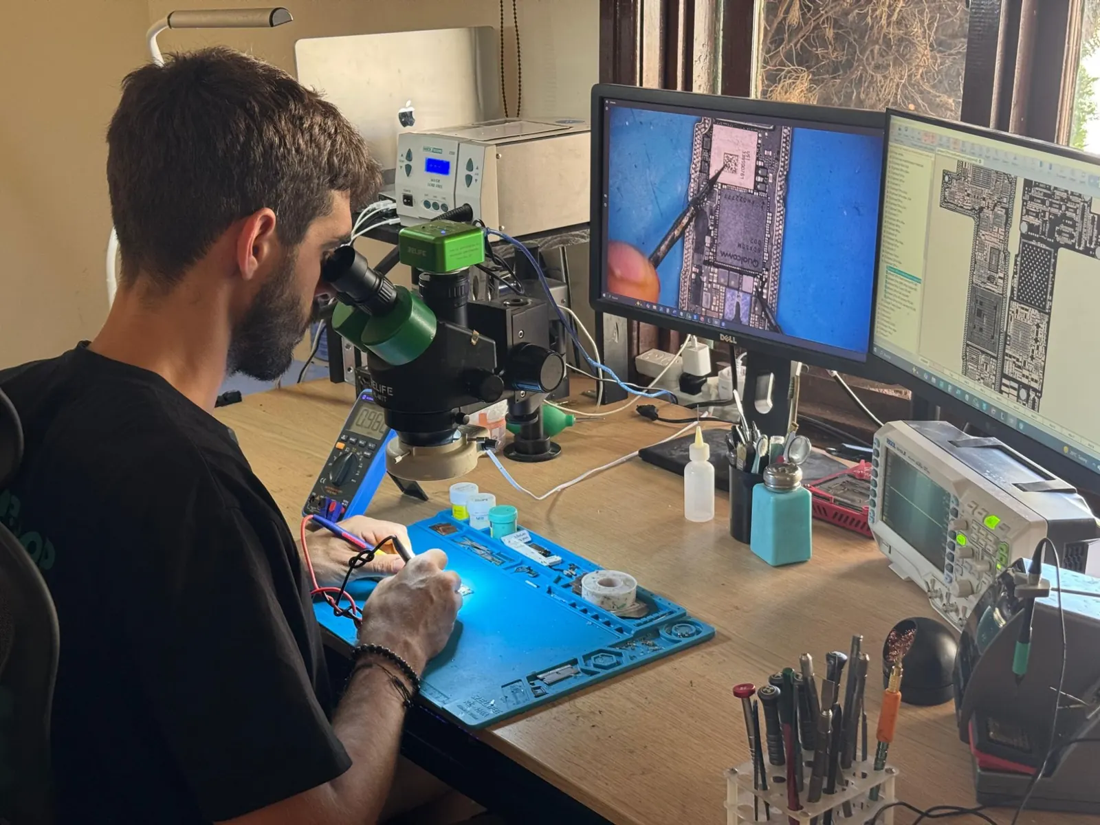 Técnico reparando una placa con microscopio, con la placa ampliada en el monitor
